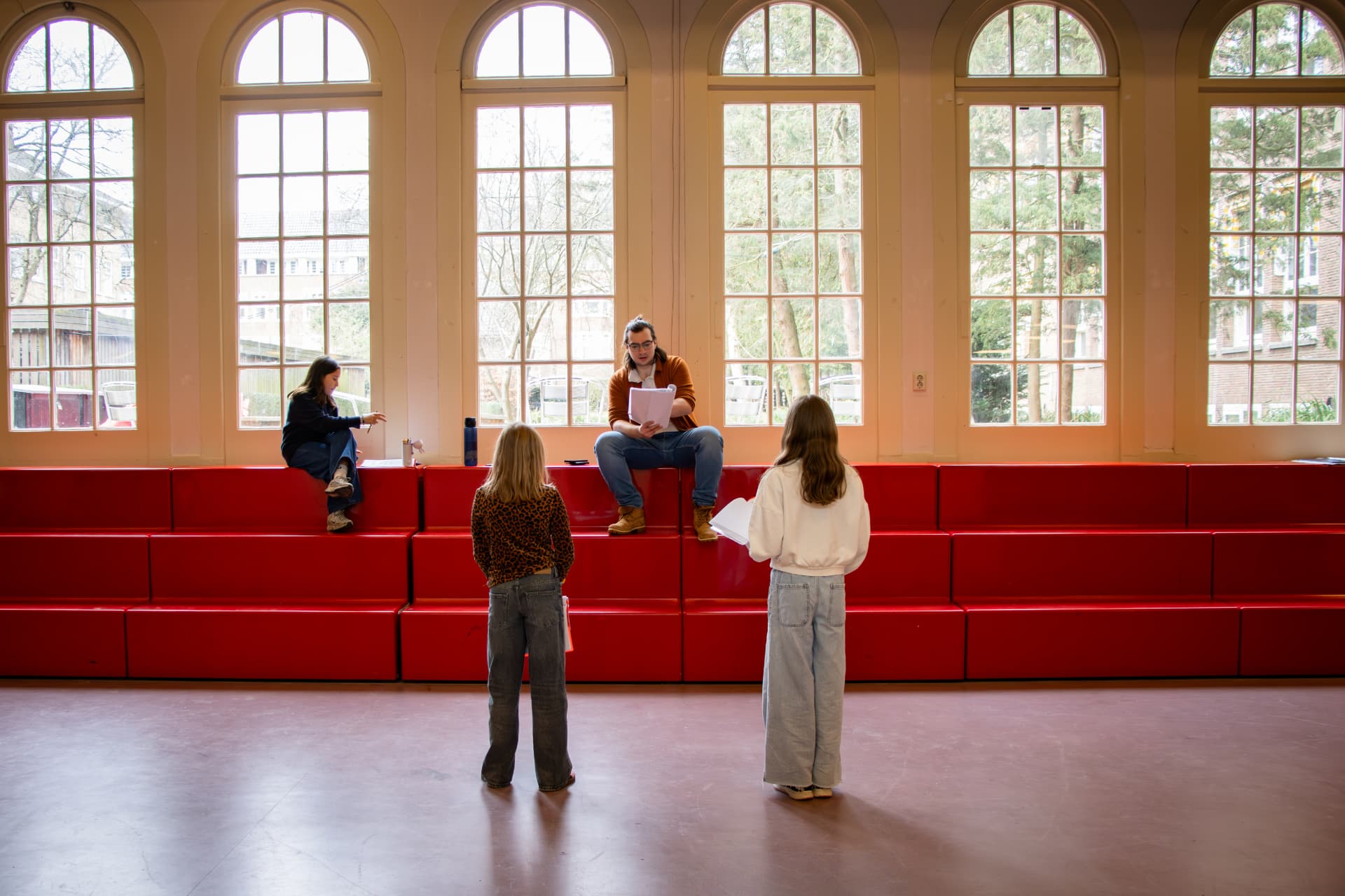 Twee leerlingen repeteren met hun docent bij de rode tribune in de aula